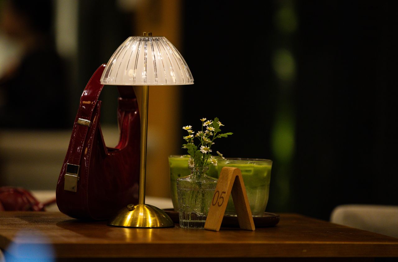 about-us-02 A warm cafe table setting with a lamp, handbag, green beverage, and flowers.
